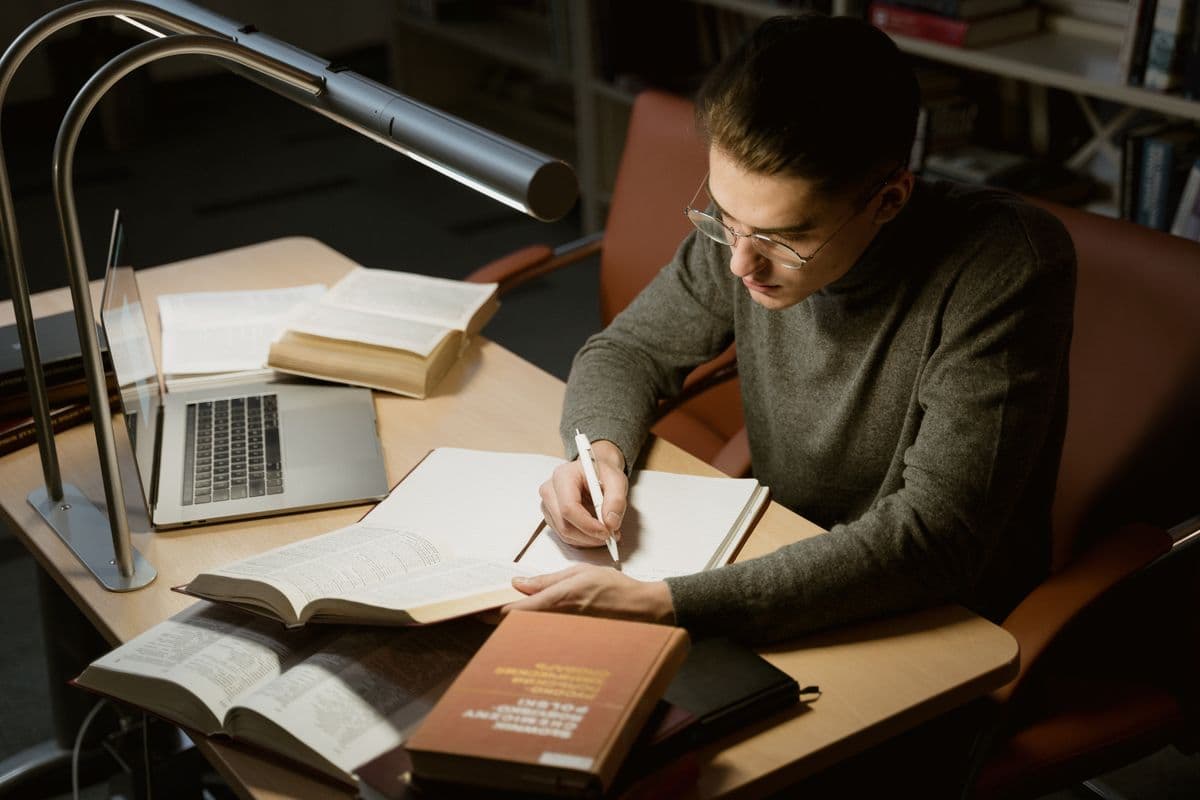 Студент в очках вечером пишет в тетради за столом библиотеки рядом с книгами, ноутбуком и настольной лампой