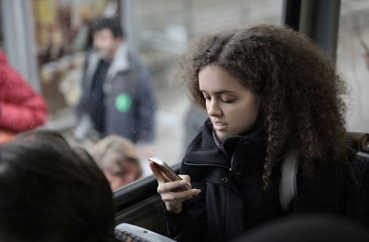 Кудрявая студентка с рюкзаком сидит у окна в автобусе и смотрит в смартфон