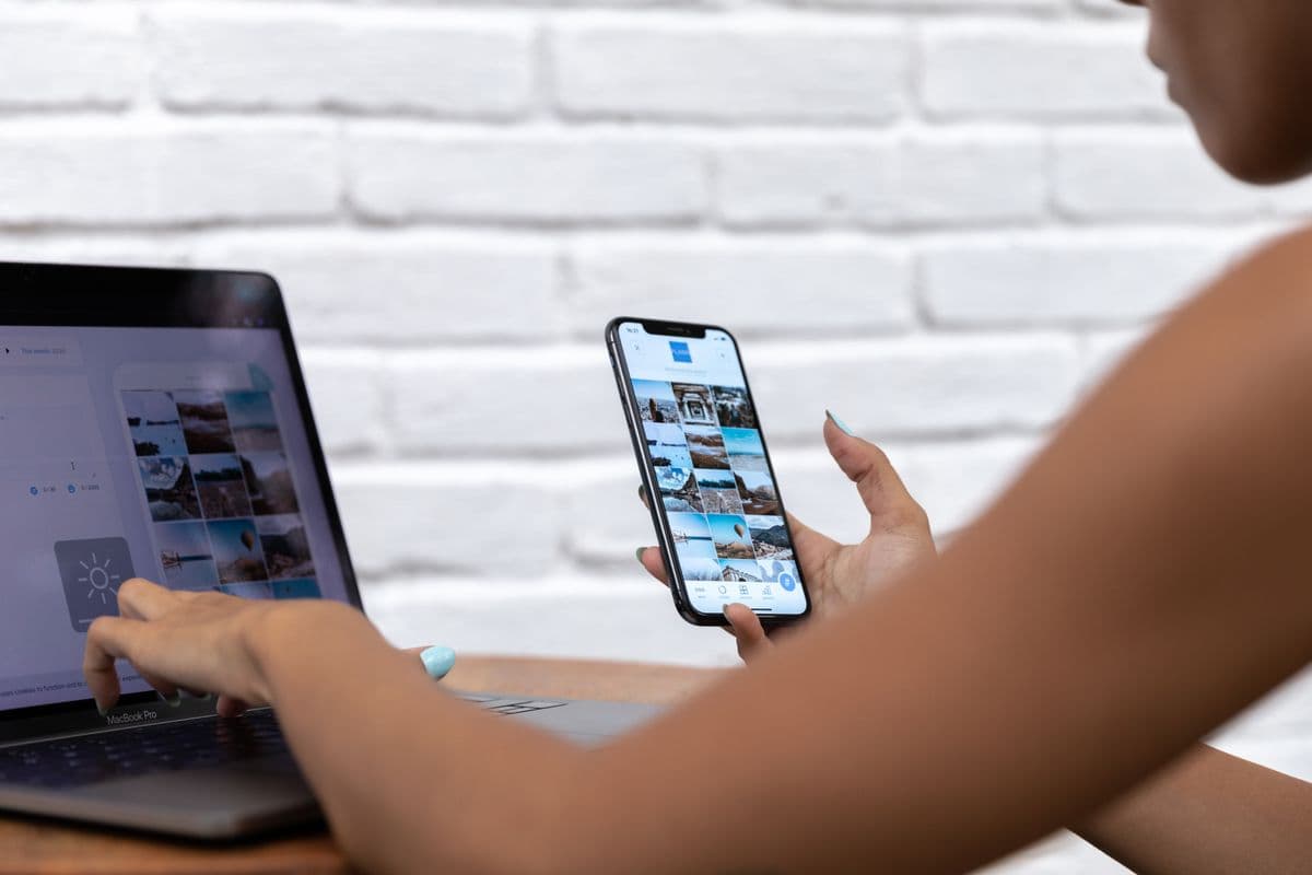 Девушка у белой кирпичной стены печатает на ноутбуке и держит смартфон с лентой соцсети