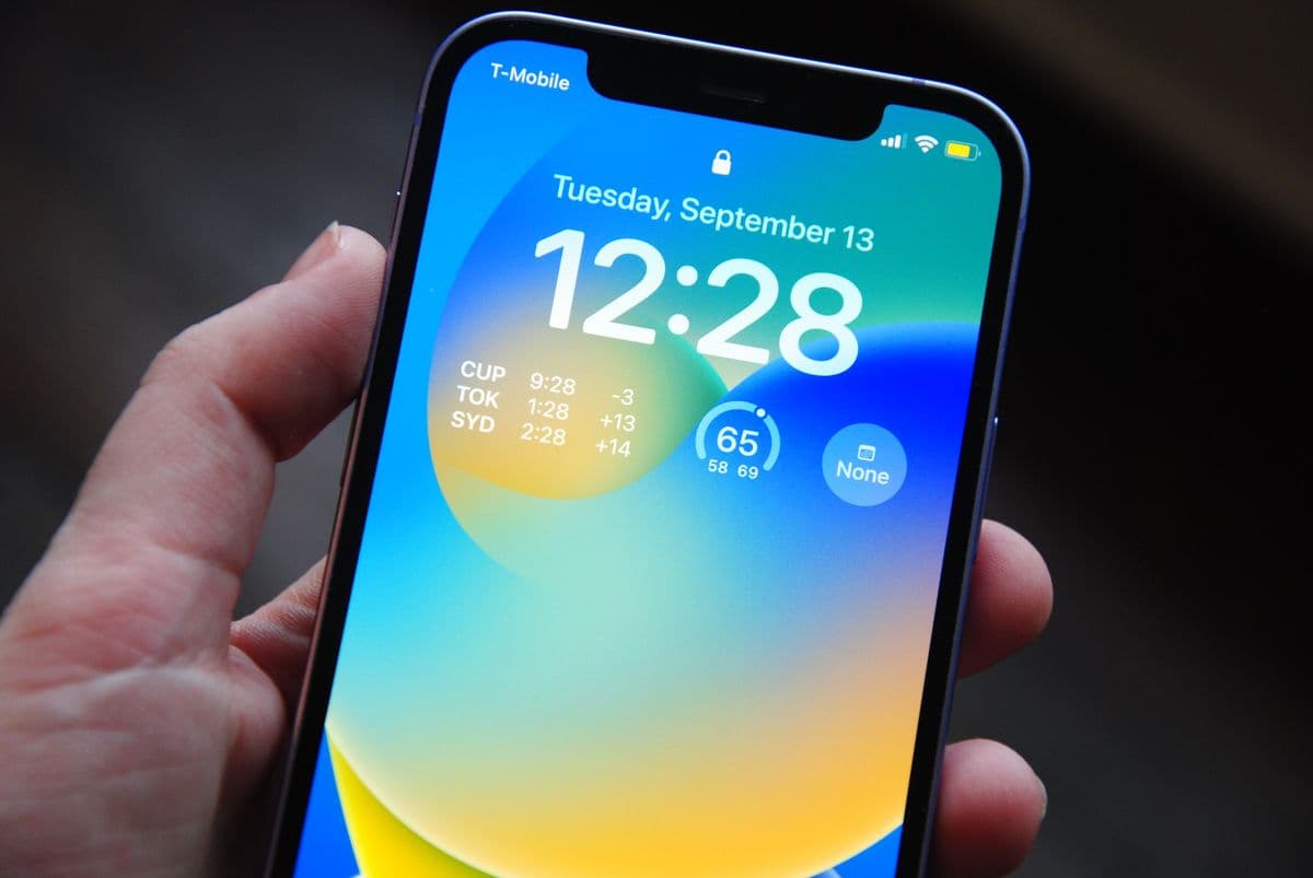 Рука держит iPhone: на экране блокировки сине-жёлтые обои, время, дата и виджеты