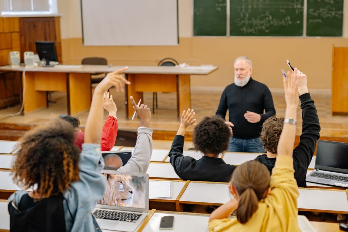Студенты с ноутбуками в аудитории поднимают руки перед преподавателем у зелёной доски