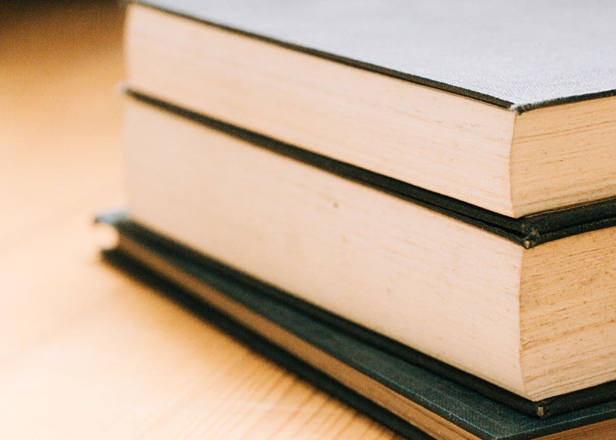 Две толстые книги в тёмном твёрдом переплёте лежат стопкой на светлом деревянном столе