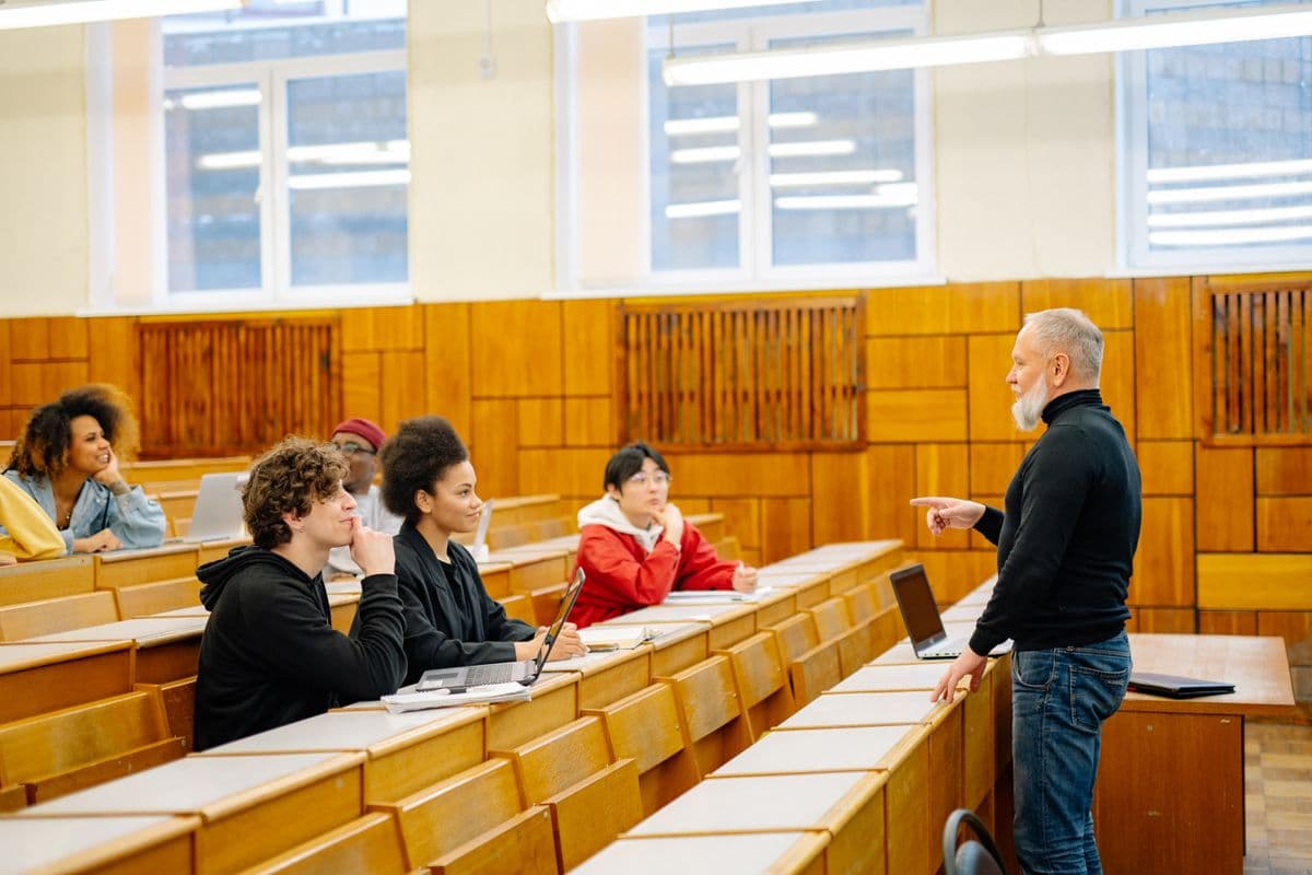 Седобородый преподаватель в чёрной водолазке объясняет материал студентам с ноутбуками в лекционной аудитории