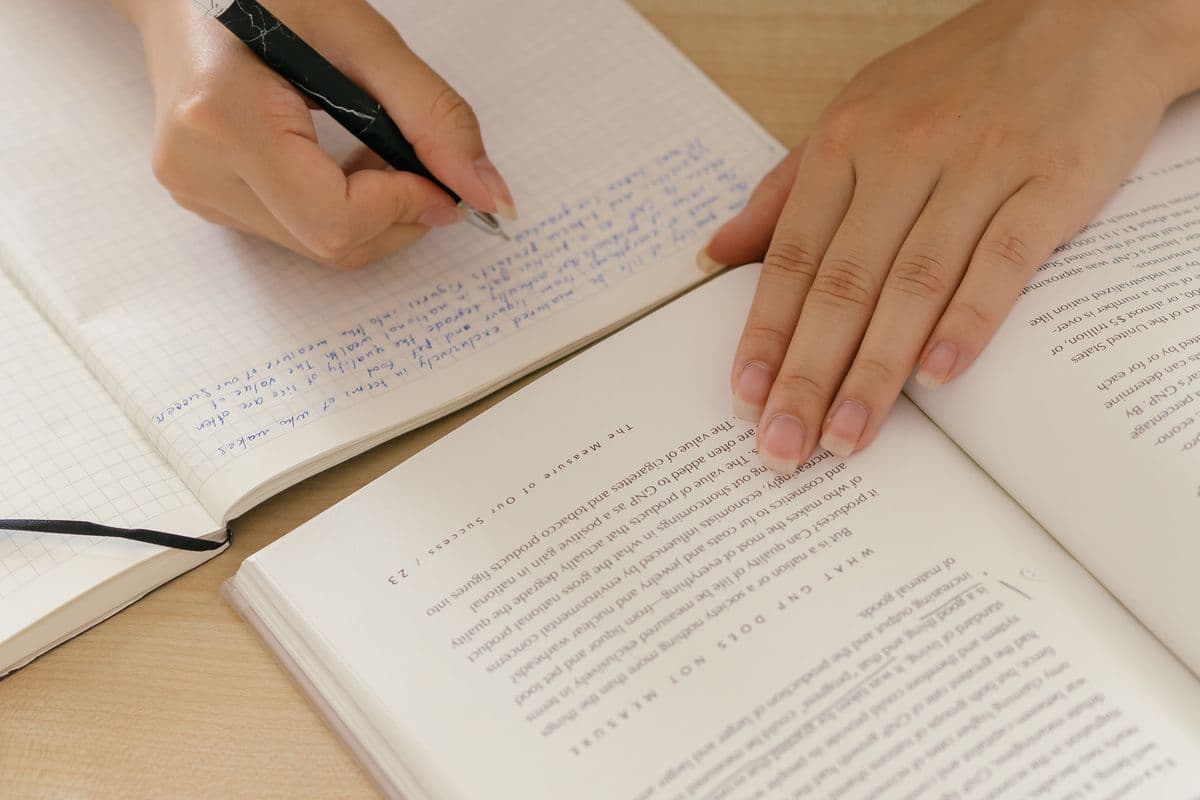 Студент держит открытую книгу и пишет своими словами конспект в тетради в клетку
